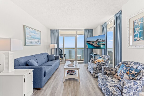 A living room with a sofa, a coffee table, and a patterned armchair. There is a view of the sea through the balcony doors.