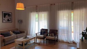 Un salon avec un canapé, des fauteuils, une table basse et une lampe. Il y a des rideaux aux fenêtres et un tableau encadré au mur.