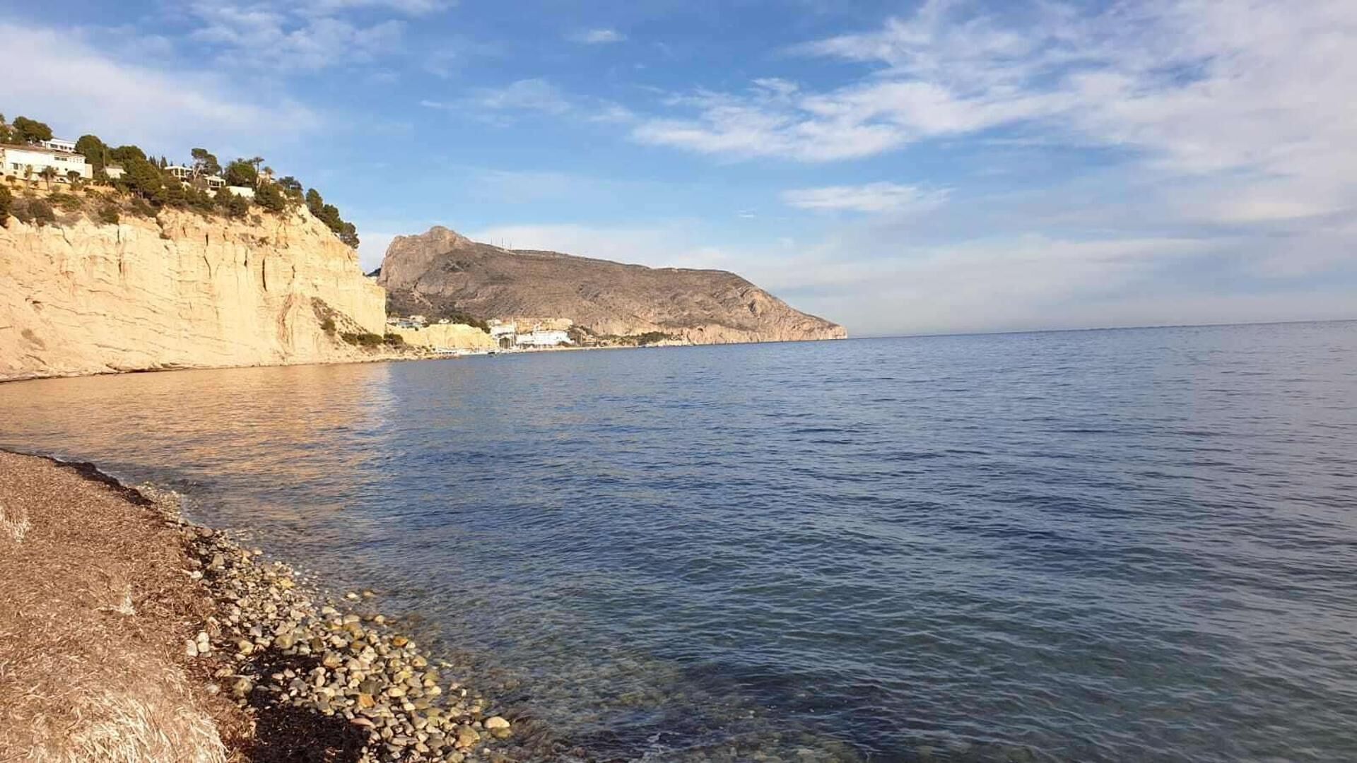 Escena costanera amb una platja rocosa, aigua cristal·lina i un penya-segat gran amb vegetació escassa.