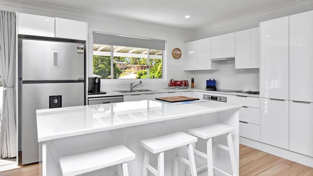 A modern kitchen with white cabinetry, a central island with stools, a coffee maker, and a view of greenery outside.