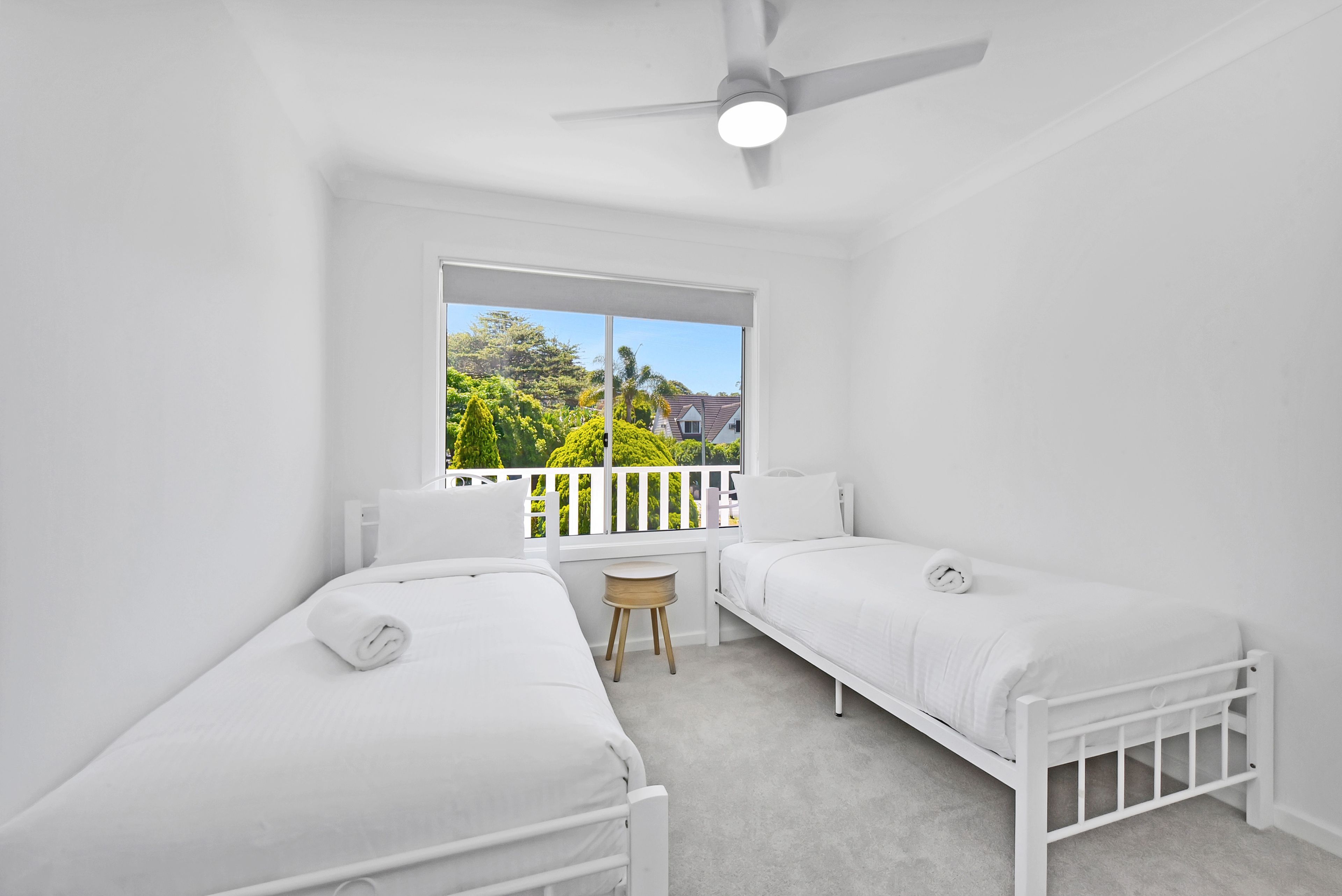 A bedroom with two single beds, a ceiling fan, a window with a view of greenery, and a small table.