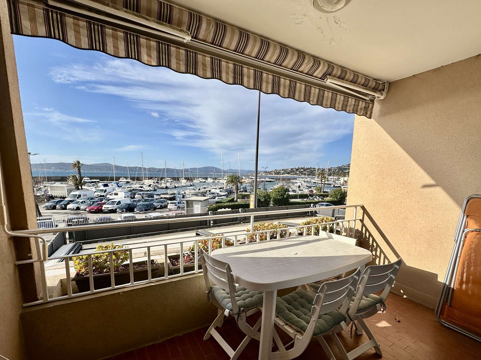 A balcony with a table and chairs, a view of a marina with boats, and a parking lot.