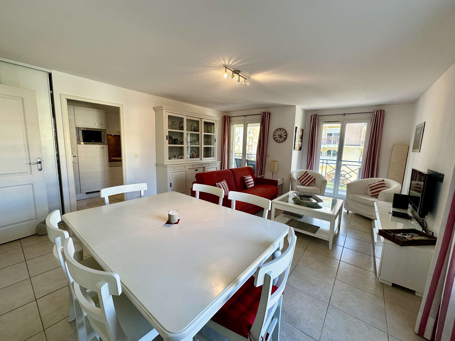 A well-lit living area with a dining table, red and white sofas, a TV, and a kitchen area with cabinets and a microwave.