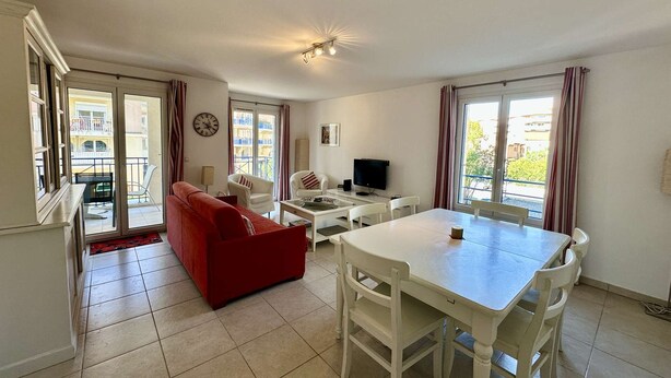 A living room with a red sofa, white armchairs, a dining table with chairs, a TV, and a balcony with curtains. - APARTMENT T3 AIR-CONDITIONED FOR RENT FOR 4 PEOPLE IN THE CITY CENTER (STE MAXIME)