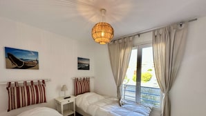 A bedroom with two beds, a nightstand, a lamp, and a hanging light fixture. There are two paintings on the wall and a window with curtains. - APARTMENT T3 AIR-CONDITIONED FOR RENT FOR 4 PEOPLE IN THE CITY CENTER (STE MAXIME)