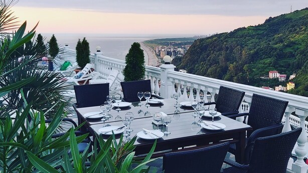A dining table set for six with chairs, overlooking a coastal cityscape and hills. - Hotel zurapalace (Batumi)