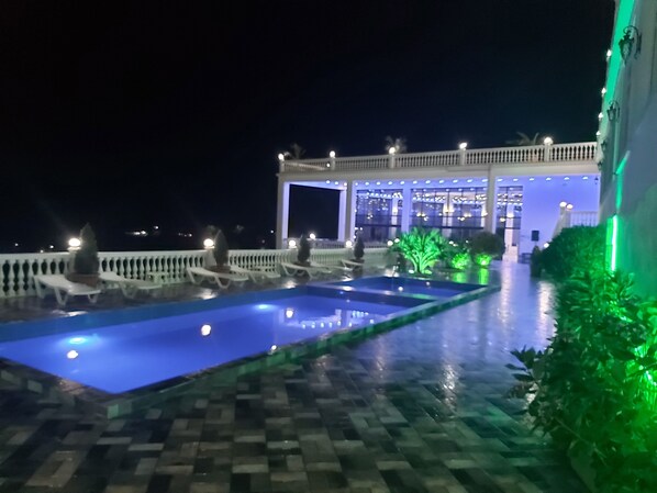 Une vue nocturne d’un hôtel doté d’une piscine, d’un espace couvert aménagé avec des sièges et d’une entrée bien éclairée.