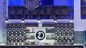 A bar area with a chandelier, shelves of bottles, and a logo. - Hotel zurapalace (Batumi)