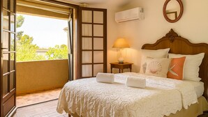 A hotel room with a bed, a nightstand with a lamp, a wall-mounted air conditioner, and a balcony with a view of greenery. - Villa Caline | Wi-Fi, Clim, 150 m from the beach (Lucciana)