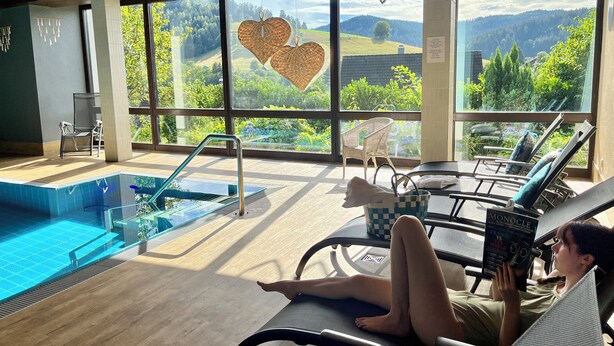 A poolside area with a view of a mountainous landscape, a lounge chair, and a person reading a book. - Apart Hotel Goldener Hahn Baiersbronn, Ferienwohnungen | Frühstück, Sauna & Indoorpool (Baiersbronn)