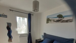 A bedroom with a bed, a nightstand, a lamp, a window with curtains, a wall-mounted air conditioning unit, and a painting. - Under the Alpilles sun  (Mouriès)