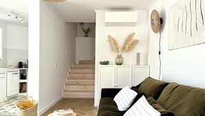 Una sala moderna con un sofá marrón, un taburete de madera y un tapete verde. Una escalera conduce a una zona de cocina con armarios blancos y un ventilador colgante.