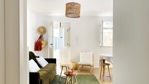 Una sala de estar acogedora con un tapete verde, un sofá con almohadas a rayas, un sillón de mimbre, una pequeña mesa de madera con una taza, y una cesta colgante.