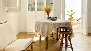 Un comedor con una mesa cubierta por un mantel a cuadros, dos taburetes de madera y una silla blanca acolchada. Se observa un aparador de cristal con vajilla y un jarrón con flores secas.