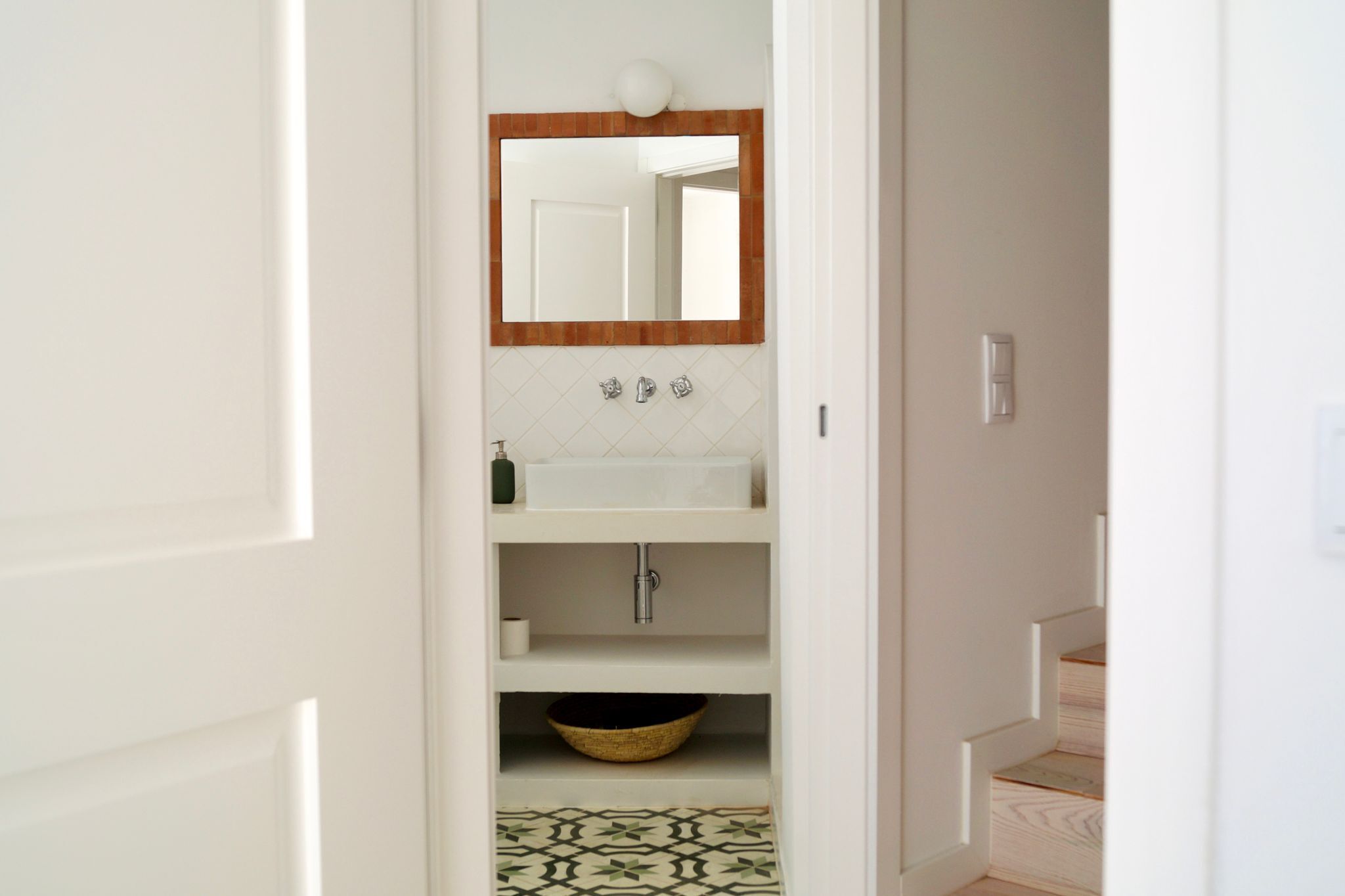 Une salle de bain avec un lavabo blanc, un miroir avec un cadre en bois et un carrelage décoré.