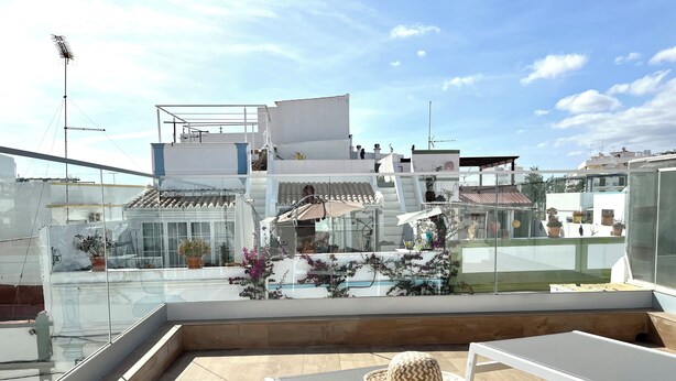 A rooftop terrace with a glass balustrade, a white lounge chair, a wooden tray with croissants and coffee cups, and potted plants.