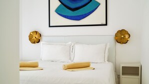 A neatly made bed with white linens and a rolled-up towel, a bedside table with a lamp, and a framed abstract painting above the bed. - Casa Pasha - Olhão, Algarve (Olhão)