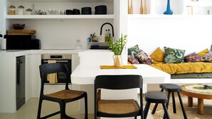 A modern kitchen with white cabinetry, a dining area with a white table and black chairs, and a living area with a sofa and cushions. - Casa Pasha - Olhão, Algarve (Olhão)