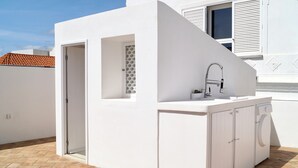 A compact outdoor kitchen with a sink, faucet, and washing machine, set against a white wall with a tiled roof visible in the background. - Casa Pasha - Olhão, Algarve (Olhão)