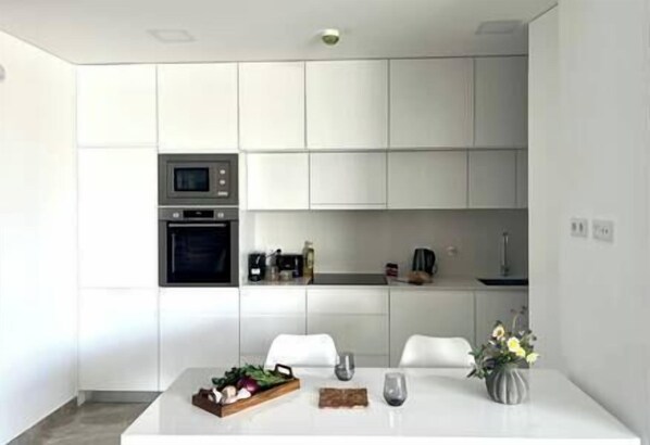 A modern kitchen with white cabinetry, a built-in oven, and a microwave. There is a dining table with a vase of flowers and a tray of vegetables.