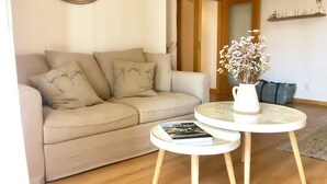 A living room with a beige sofa, a round glass table with a book, a clock on the wall, and a small potted plant. - Luz Marina - Olhão, Algarve (Olhão)