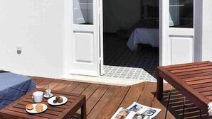 Una terraza de madera con una mesa lista para el desayuno, una cama visible a través de una puerta abierta y una pequeña alfombra en el suelo.