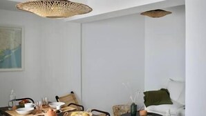 Un área de comedor con una mesa de madera dispuesta para dos personas, una lámpara de fibra trenzada colgando arriba, y un cojín verde en un sofá al fondo.