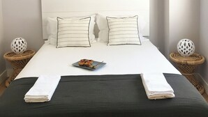 A neatly made bed with two pillows, a bedside table with a decorative item, and a wall-mounted headboard. - Luz Cubo - Olhão, Algarve (Olhão)