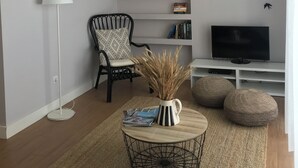 A living room with a black chair, white lamp, TV, bookshelf, and decorative wall art. - Luz Cubo - Olhão, Algarve (Olhão)