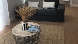 A modern living room with a black sofa, a round coffee table with magazines, and a kitchen in the background with a refrigerator, microwave, and sink. - Luz Cubo - Olhão, Algarve (Olhão)