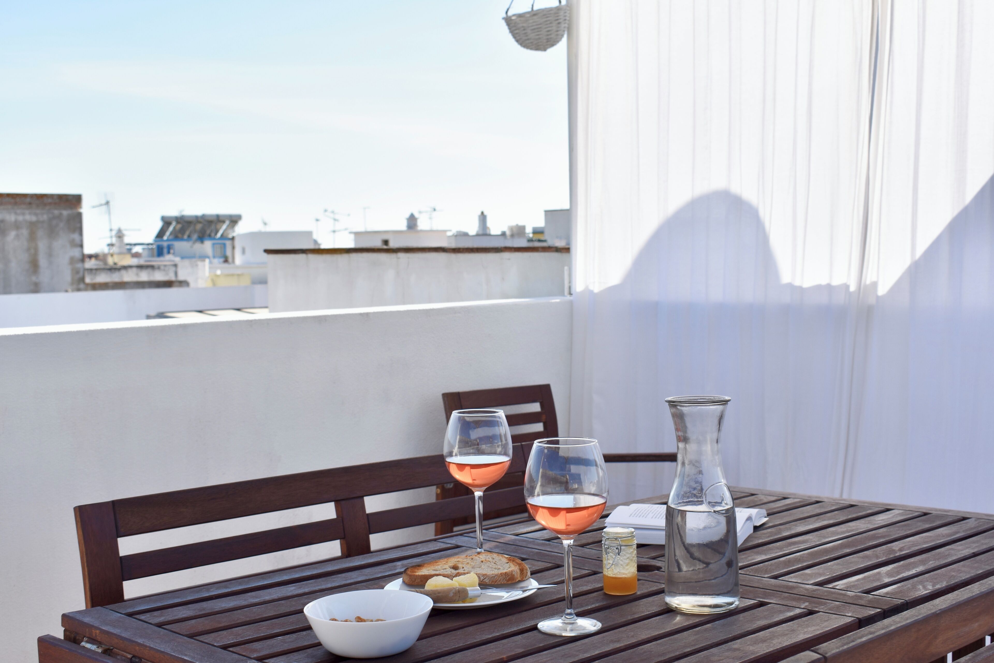 A wooden table with a white bowl, two glasses of rosé wine, a bottle of water, and a small jar on a balcony overlooking a cityscape.