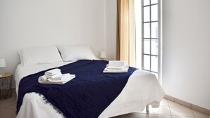 A neatly made bed with white linens and a dark blue blanket, a small bedside table with a lamp, and a window with curtains. - Luz Santana - Olhão, Algarve (Olhão)