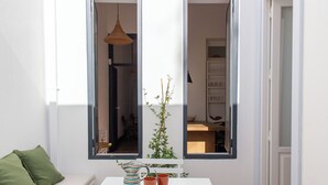 Huoneessa on valkoinen pöytä, vihreä koristetyyny, raidallinen kannu ja kaksi oranssia kuppia puisen alustan päällä.