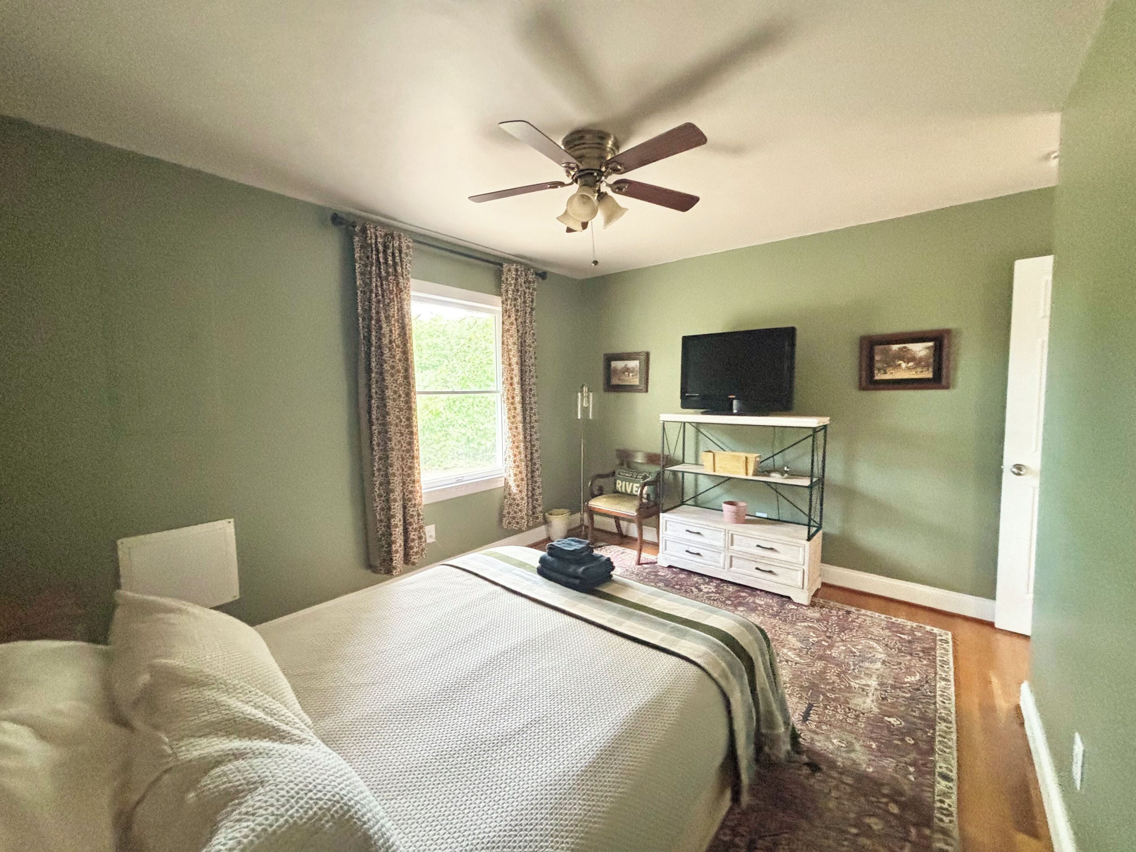 A bedroom with a bed, a ceiling fan, a window with curtains, a television on a shelf, and a door.