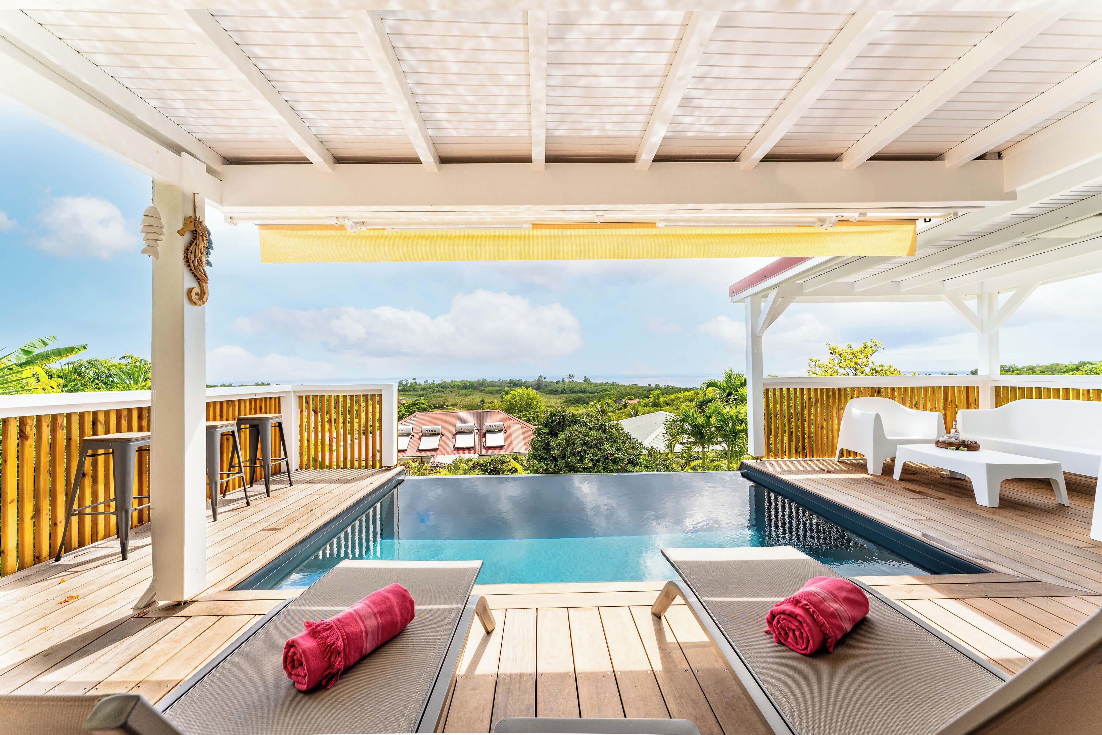 A rooftop infinity pool with wooden deck, white lounge chairs, and a view of the landscape.