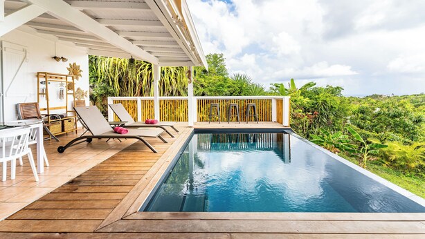 A pool area with a wooden deck, lounge chairs, and a view of lush greenery.