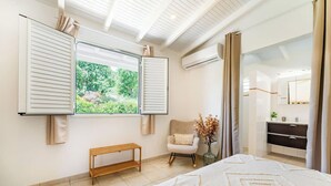A bedroom with a large bed, a window with shutters, a chair, a bench, and a dresser with a mirror.