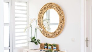 A modern interior with a white door, a round mirror with a woven frame, a wooden console table with potted plants, and a tiled floor.