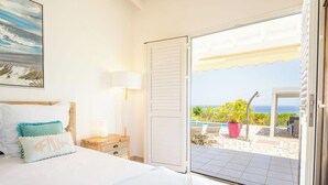 A bedroom with a bed, a nightstand, a lamp, and a view of an outdoor patio with a pool and plants.