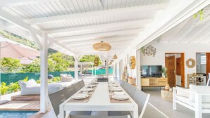 Un espacio de comedor al aire libre cubierto, con piscina, mobiliario blanco y un televisor al fondo.