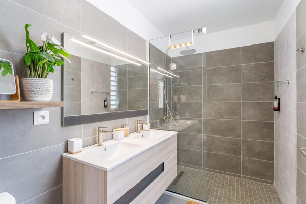 A modern bathroom with a double sink vanity, a large mirror, a potted plant, and a glass-enclosed shower area.