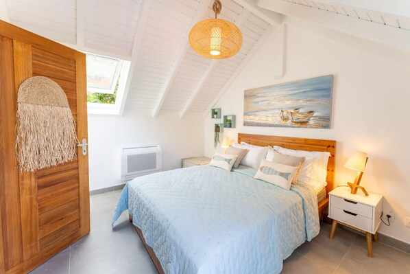 Un dormitorio con una cama grande, cabecera de madera, mesitas de noche con lámparas, un cuadro de un barco en la pared y una ventana con vista a la vegetación.