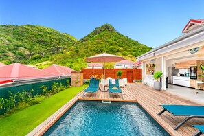A swimming pool with lounge chairs and umbrellas, a wooden deck, and a kitchen area.