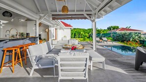 Un espacio exterior techado con piscina, mobiliario blanco y una zona de cocina con barra.