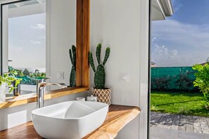 A modern bathroom with a large white sink, wooden countertop, and a view of a green garden through a glass door. - Villa Paloma with a private pool and 2 air-conditioned bedrooms (Grand-Bourg)