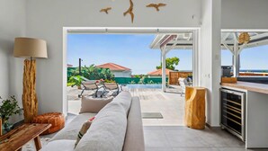 Una sala moderna con un sofá, una mesa de centro de madera y una lámpara. Se ve un patio con mobiliario exterior y una piscina.