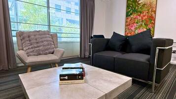 A modern hotel room with a large window, a sofa, a chair, and a coffee table with books.