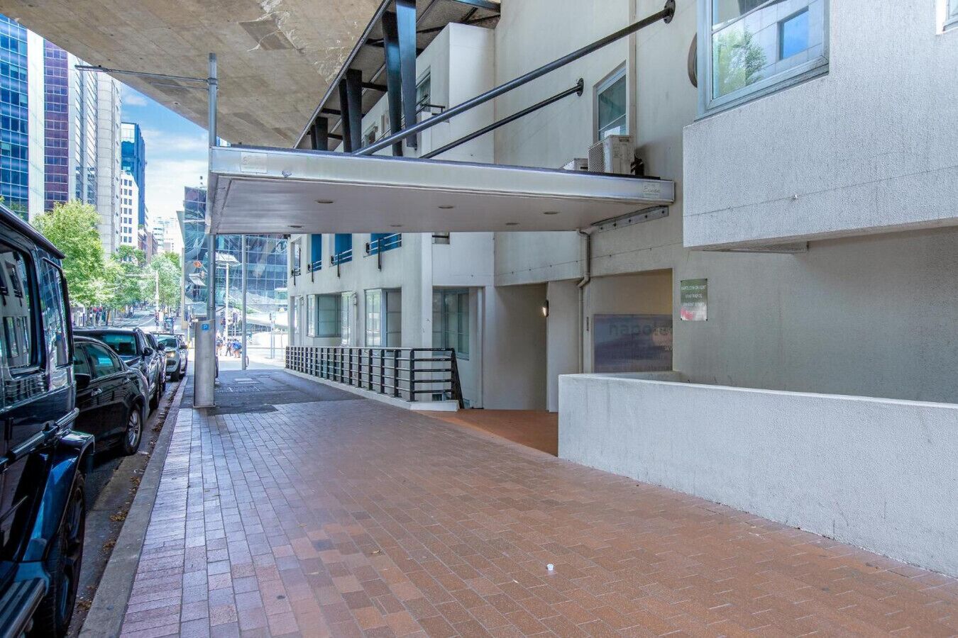 A covered pedestrian walkway with parked cars on one side and a building with a white facade and large windows.