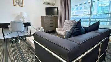 A modern hotel room with a sofa, a desk, a chair, a dresser, and a large window with a city view.