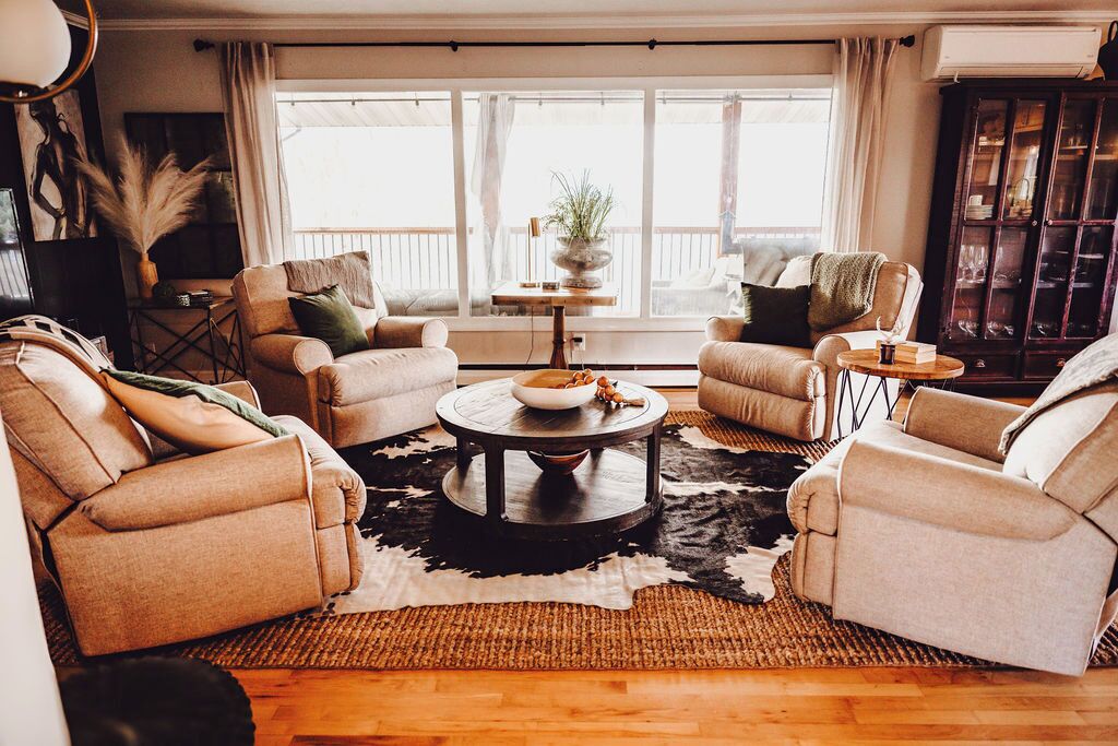 Ein gemütliches Wohnzimmer mit zwei beigefarbenen Sofas, einem runden Couchtisch und einem großen Fenster mit Vorhängen.